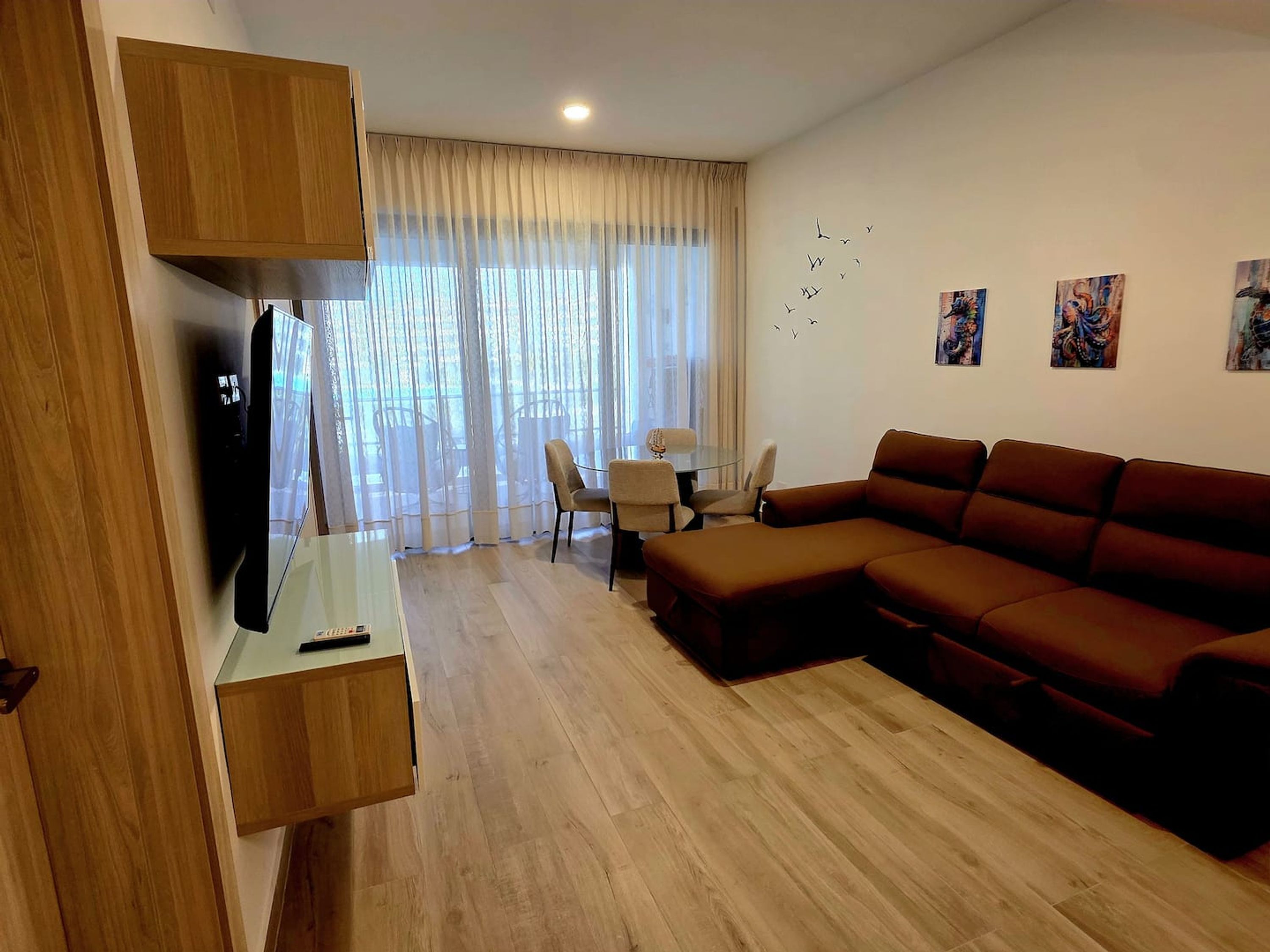 A modern living room with a sectional sofa, a glass coffee table, and a TV mounted on a wooden cabinet. There is a dining area with a table and chairs, and a large window with sheer curtains.