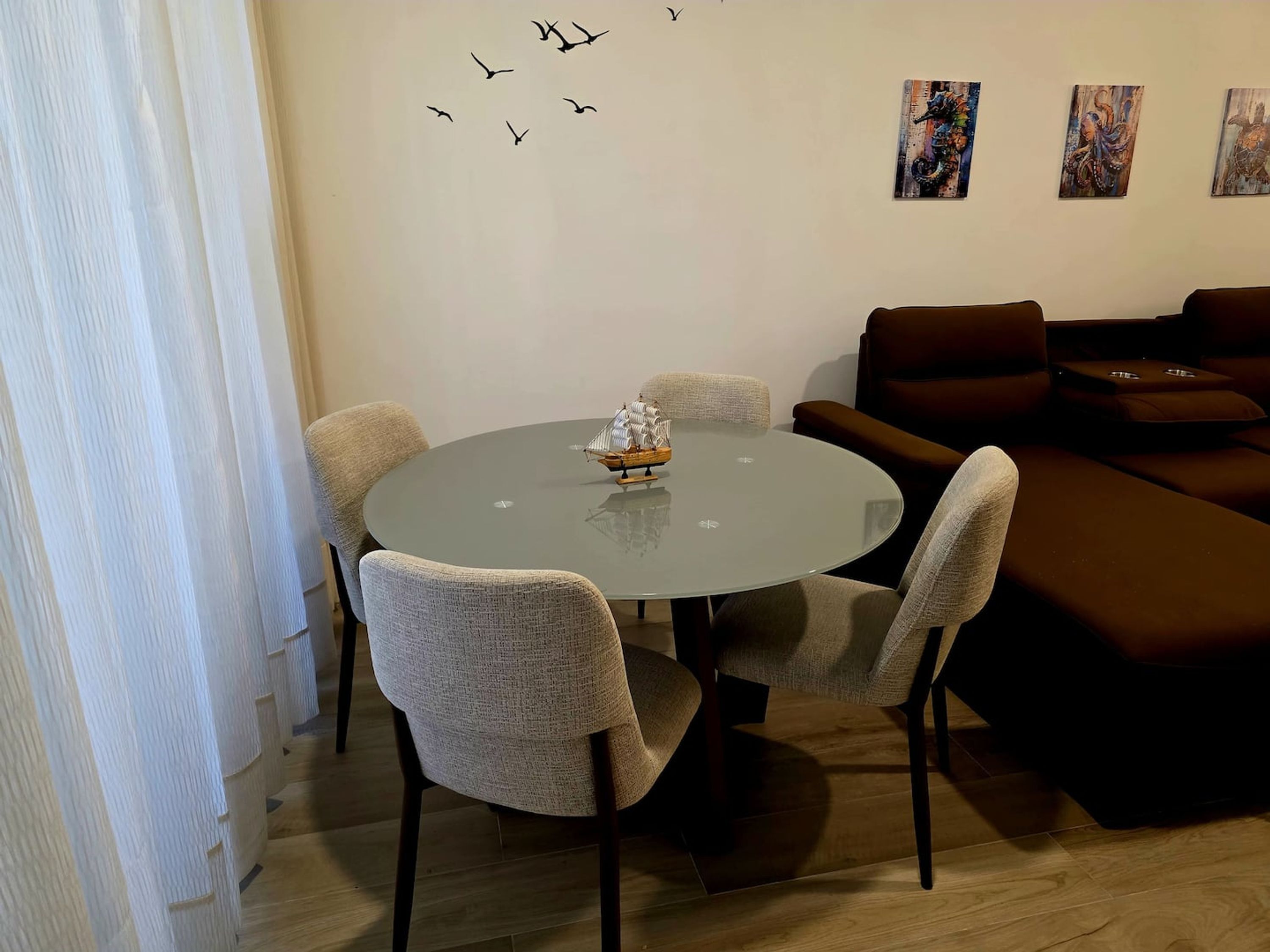 A modern living room with a round glass table, four upholstered chairs, a brown sofa, and three wall-mounted paintings.