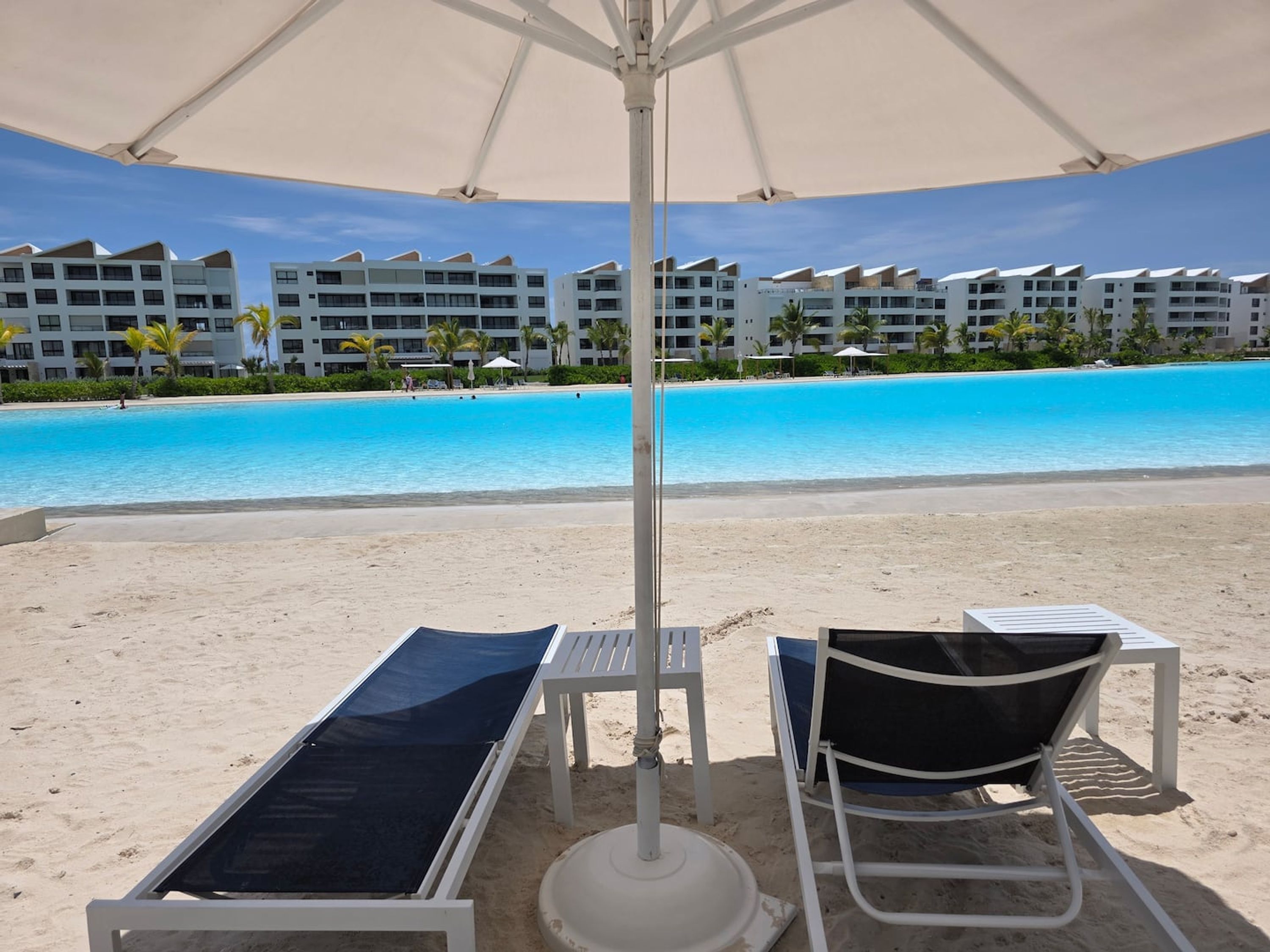 A beach with white lounge chairs and umbrellas, a clear blue pool, and a row of white buildings in the background.