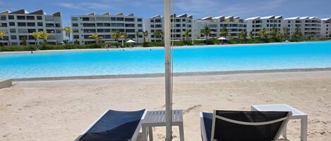 A beach with white lounge chairs and umbrellas, a clear blue pool, and a row of white buildings in the background.