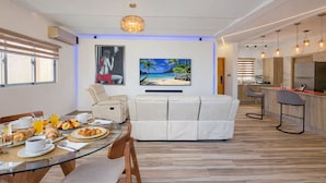 Une cuisine moderne dotée d’un îlot central, un coin repas avec une table en verre et un espace salon avec un canapé et un tableau représentant une scène de plage.