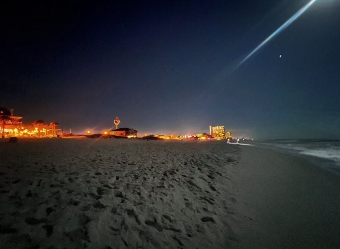 A beach at night with footprints in the sand, a building with lights, and a structure with a dome.