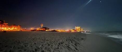 A beach at night with footprints in the sand, a building with lights, and a structure with a dome.