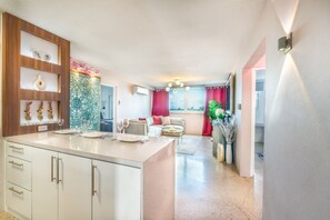 Una cocina moderna con armarios blancos y una isla central, un área de comedor con un sofá y cortinas rojas, y vista a la ciudad a través de una ventana.