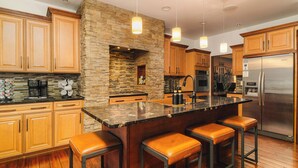 A modern kitchen with wooden cabinets, a stone accent wall, a stainless steel refrigerator, and a bar area with stools. - Lion’s Lookout | PSU Retreat w/Hot Tub & Game Room (Bellefonte)
