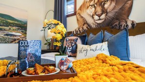 A hotel room with a large lion mural, a book titled 'Cougars on the Cliff', a tray with food, and a 'stay awhile' pillow. - Lion’s Lookout | PSU Retreat w/Hot Tub & Game Room (Bellefonte)