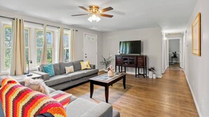 Ein geräumiges Wohnzimmer mit einem grauen Sofa, einem Couchtisch und einer bunten Decke. Eine Deckenventilator, ein Fernseher auf einem Schrank und eine Tür zu einem anderen Zimmer sind vorhanden.
