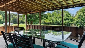 Eine überdachte Terrasse im Freien mit einem Glastisch und Stühlen, umgeben von Holzbalken und einem Metalldach, mit Blick auf eine üppige grüne Landschaft.
