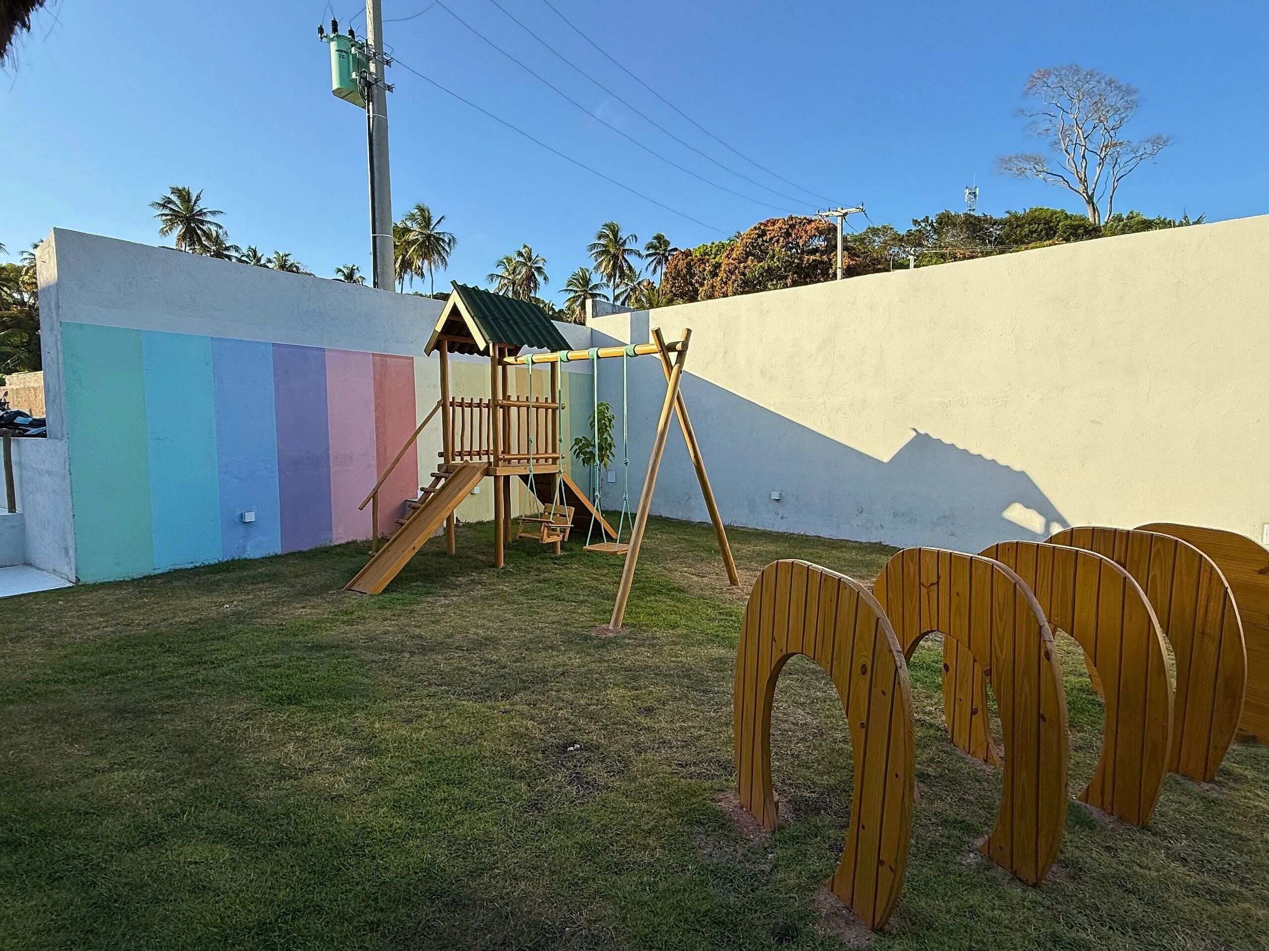 Un parque infantil con un tobogán de madera y columpios, frente a un muro con un llamativo diseño de rayas verticales.