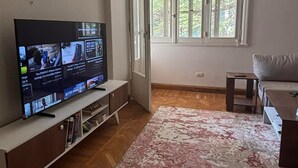 A living room with a television on a stand, a sofa, a small table, and a window with a view of greenery. - Cozy stay in prime central of Cairo, Green & Nile View. (Cairo Governorate)