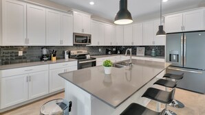Cozinha moderna com armários brancos, uma ilha central com banquetas, micro-ondas embutido e geladeira de aço inoxidável.