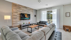 Uma sala de estar moderna com uma televisão de tela plana grande, sofás cinzas, uma mesa de centro de madeira e uma janela ampla com cortinas leves.