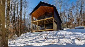 Una cabaña moderna con terraza de madera y un ventanal grande, rodeada por un paisaje nevado y árboles.