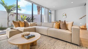 Ein modernes Wohnzimmer mit einem beigefarbenen Ecksofa, einem runden Couchtisch und einem großen Fenster mit Blick auf Palmen und eine weiße Wand.