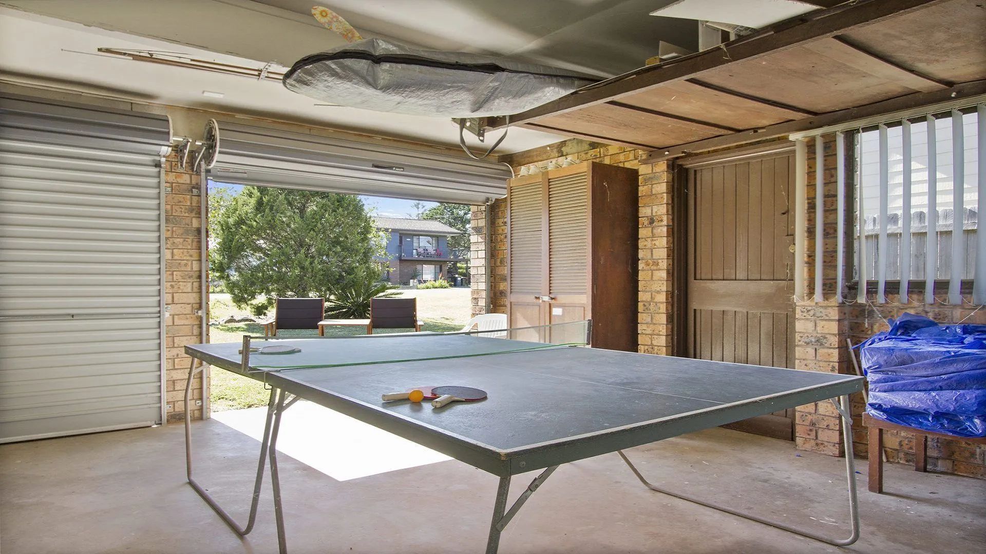 Une table de ping-pong installée dans un espace extérieur abrité, avec des murs en briques et une porte de garage.