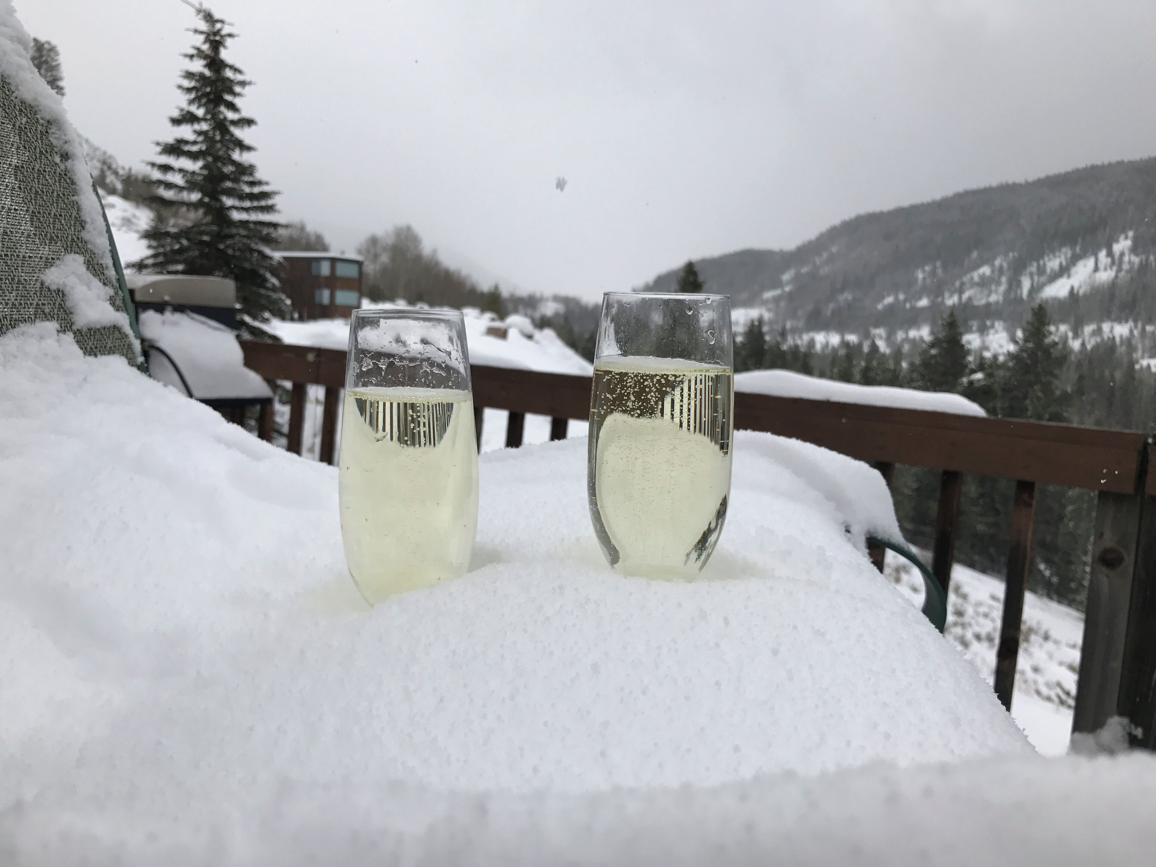 Zwei Gläser Weißwein auf einer mit Schnee bedeckten Terrasse mit Blick auf verschneite Berge im Hintergrund.