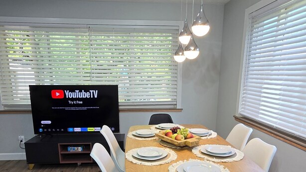 En matplats med ett träbord för fyra personer, en tv-skärm som visar YouTubeTV och ett fönster med persienner.