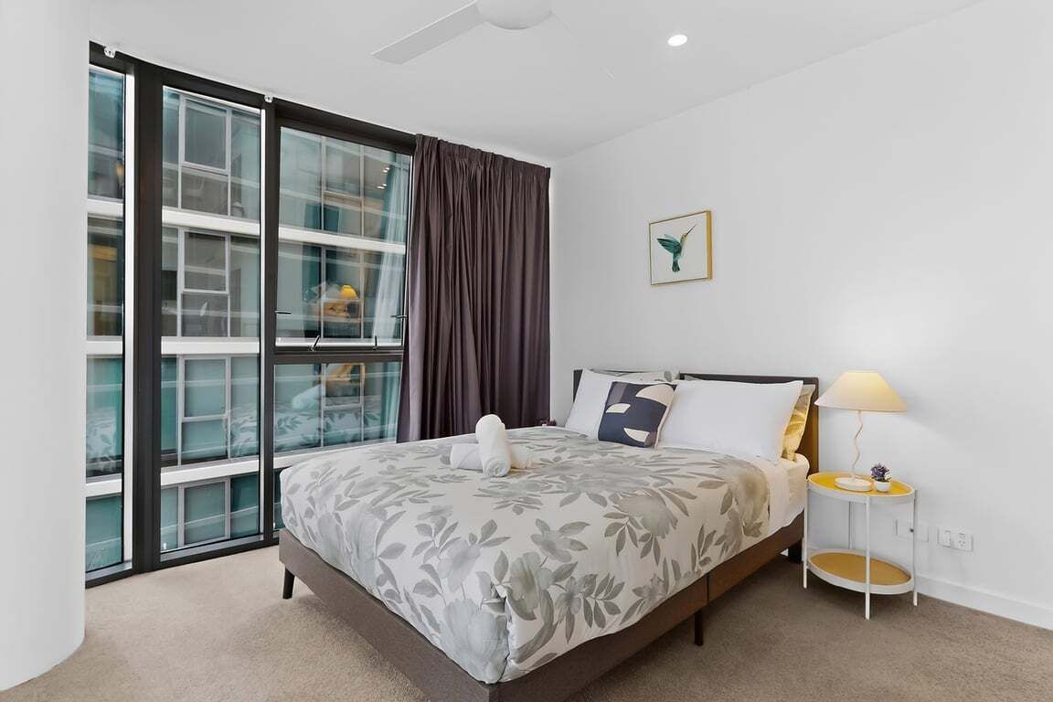 A bedroom with a bed, a window with curtains, a bedside table with a lamp, and a wall-mounted picture.
