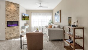 A modern living room with a stone accent wall, a flat-screen TV, a fireplace, a ceiling fan, and a glass coffee table. - Upscale 2BR/2BA Condo with Great Views, Heated Pool & Balcony (Fountain Hills)