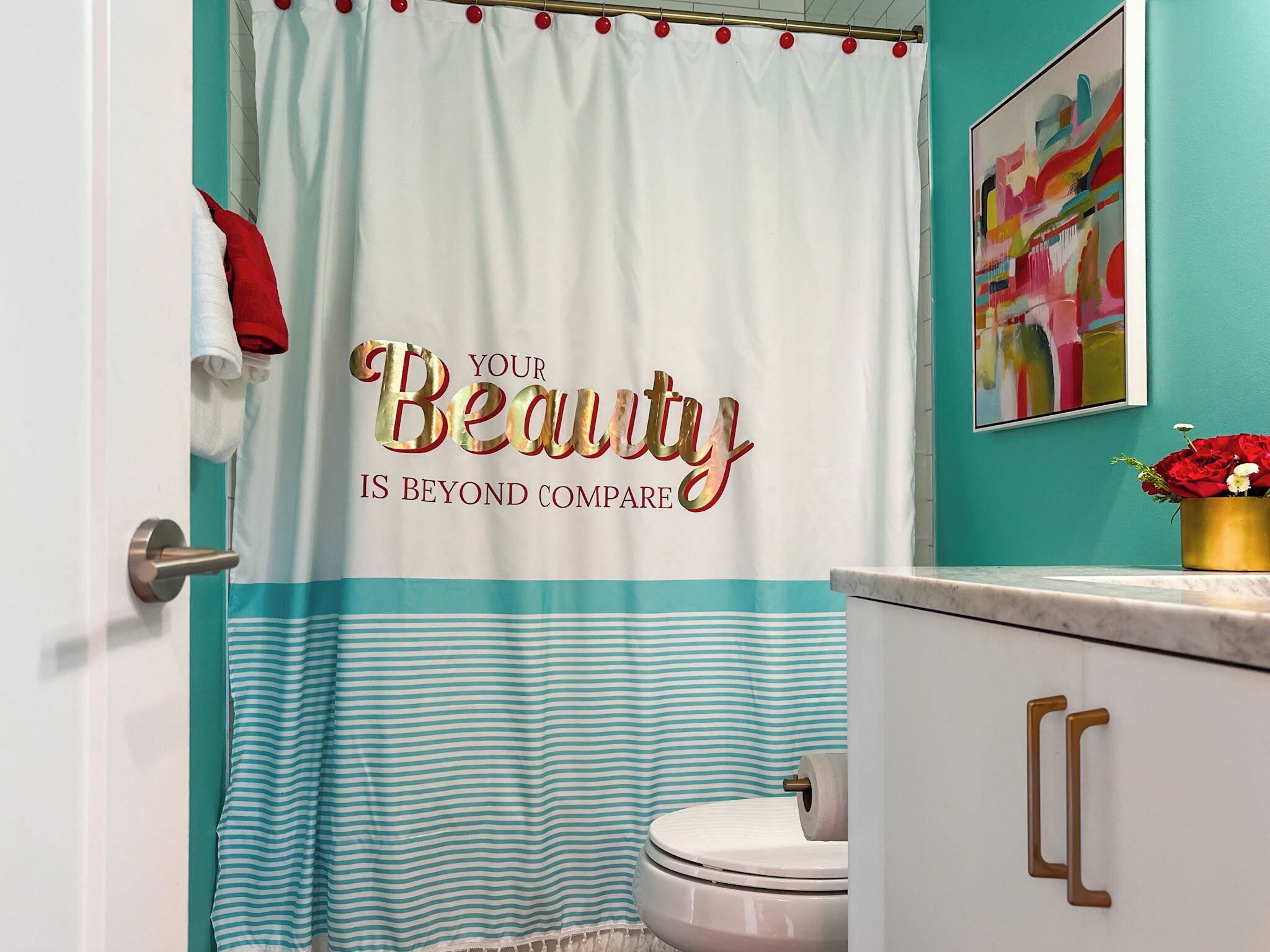 Un baño con cortina de ducha rayada en verde azulado y blanco, un inodoro, un lavabo con pomos dorados y un cuadro abstracto enmarcado en la pared.