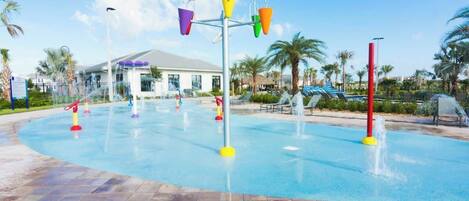 A water play area with multiple colorful water jets, a tiled walkway, palm trees, and a building in the background.