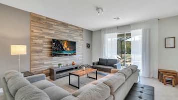 A modern living room with a large flat-screen TV, grey sofas, a wooden coffee table, and a large window with sheer curtains.