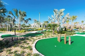 A golf course with a green putting area, palm trees, and a white building in the background. - Classy 6BR Home With Private Pool (Kissimmee)