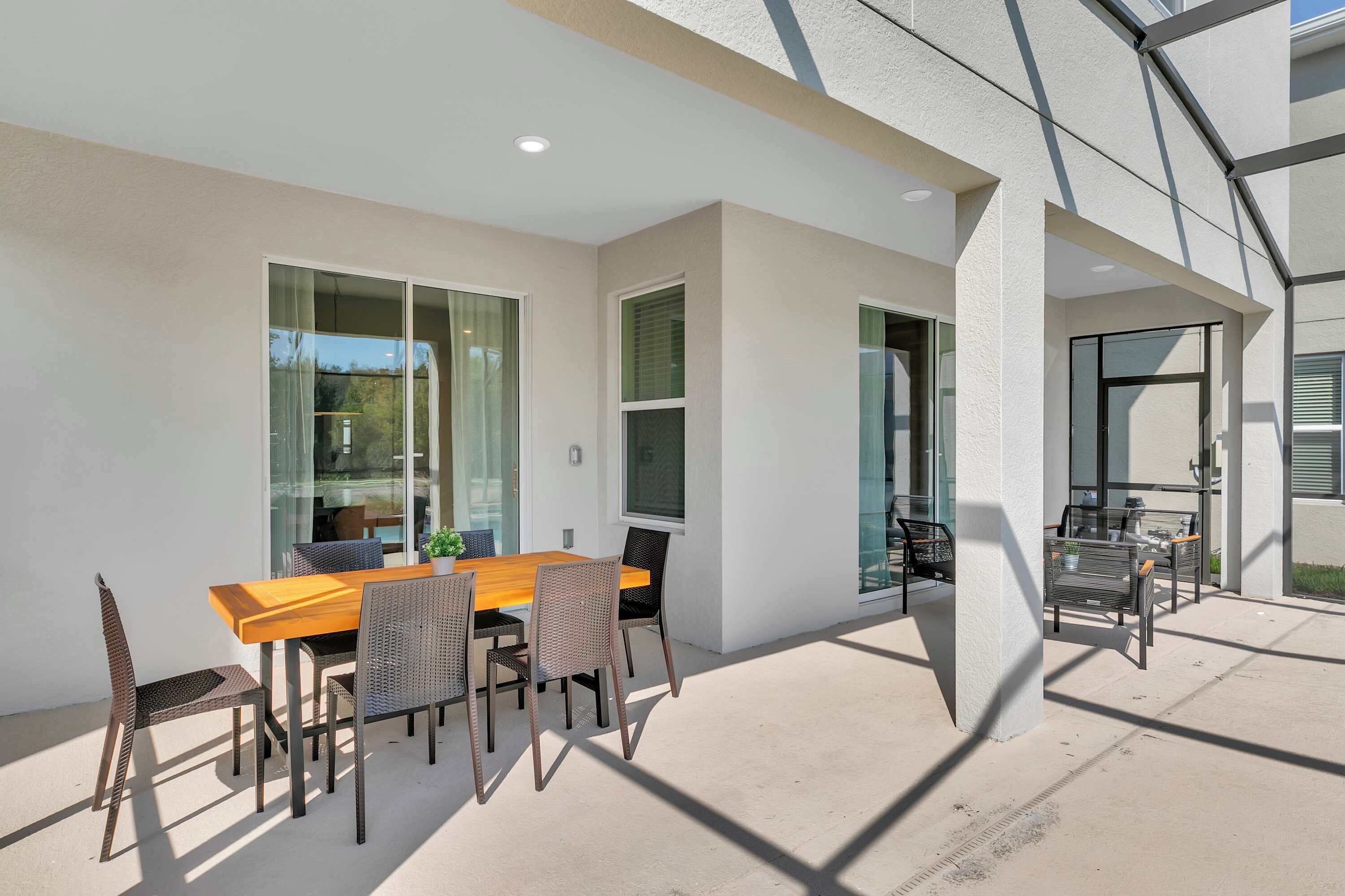 An outdoor patio area with a wooden table and wicker chairs, large windows, and a glass door leading to another space.