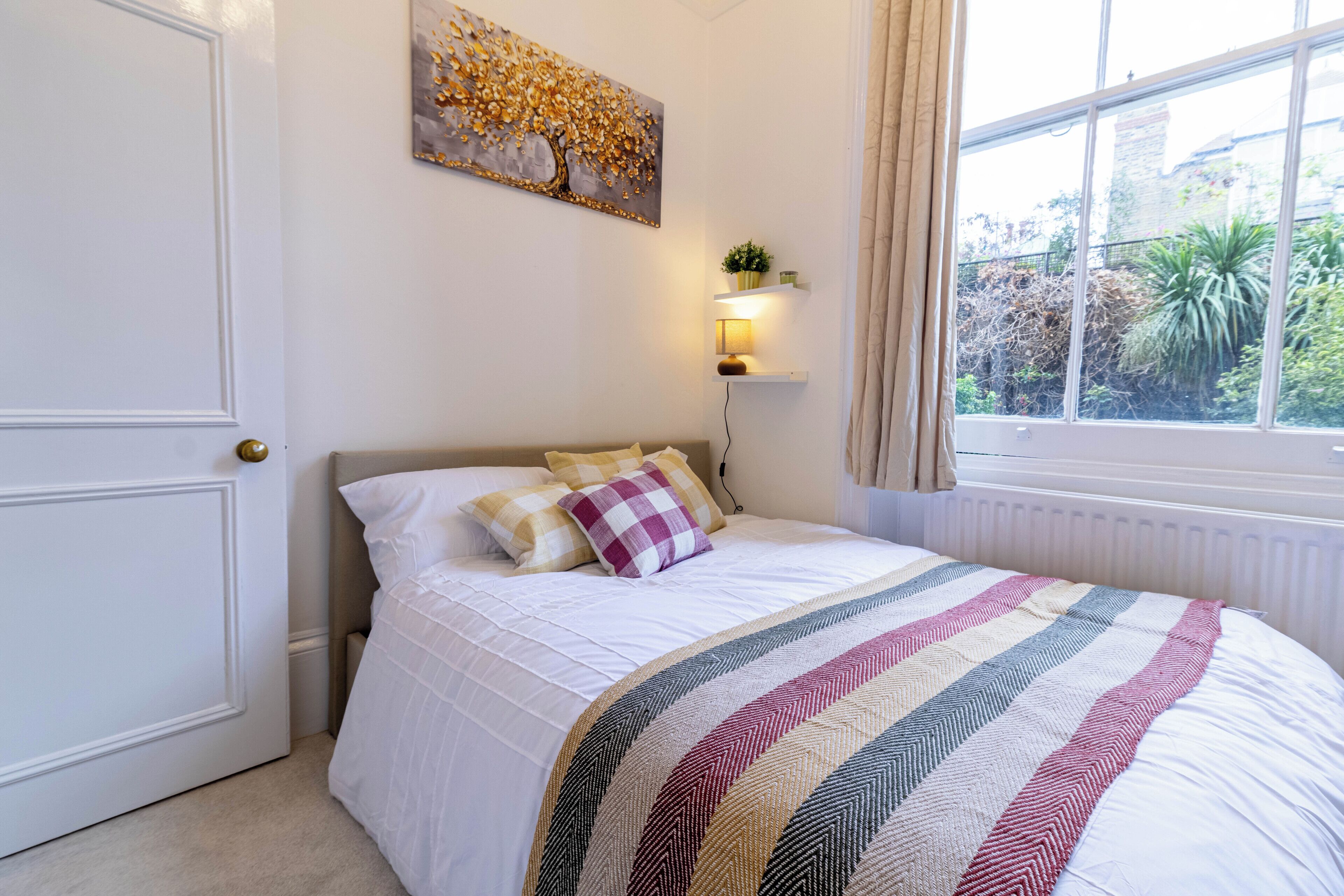 Un lit bien fait, recouvert d’une couverture à rayures, une étagère fixée au mur avec une plante, et une fenêtre donnant sur un paysage verdoyant.