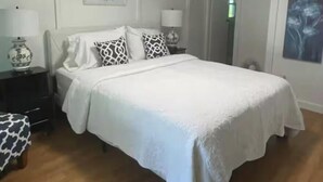 Ein Schlafzimmer mit Bett, Nachttischen, einer Lampe, einem Sessel, einer Tür und einem an der Wand hängenden Bild.