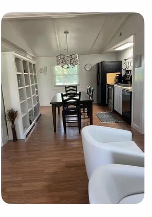 A dining area with a wooden table and chairs, a white sofa, and a kitchen with appliances. - Peaceful 3-bedroom cottage. Enjoy the Silence. (Leola)