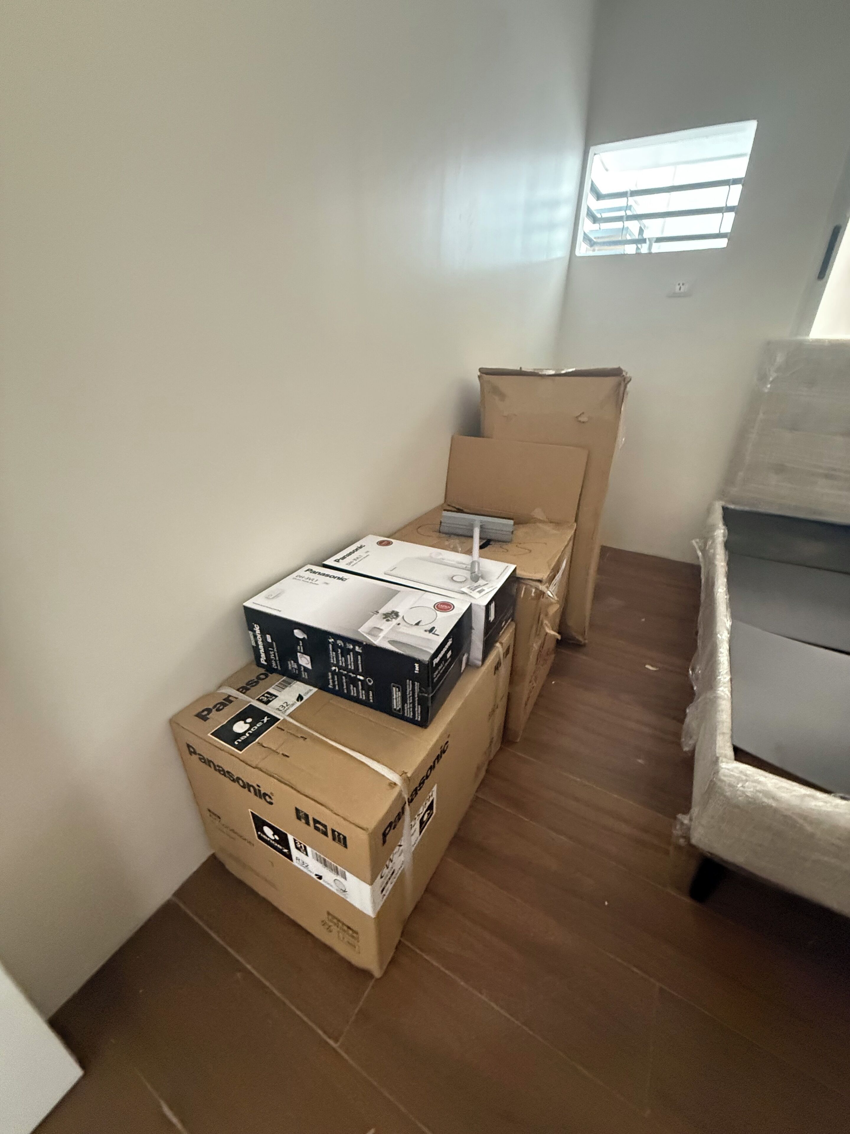 Une pièce encombrée de cartons empilés, une fenêtre avec des stores et un canapé blanc.