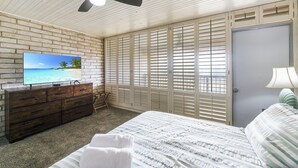 Une chambre à coucher comprenant un lit, une commode, un téléviseur, un ventilateur de plafond et une porte avec des volets.