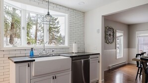 Una cocina con muebles blancos, lavavajillas de acero inoxidable, encimera negra y ventana con vistas a árboles.