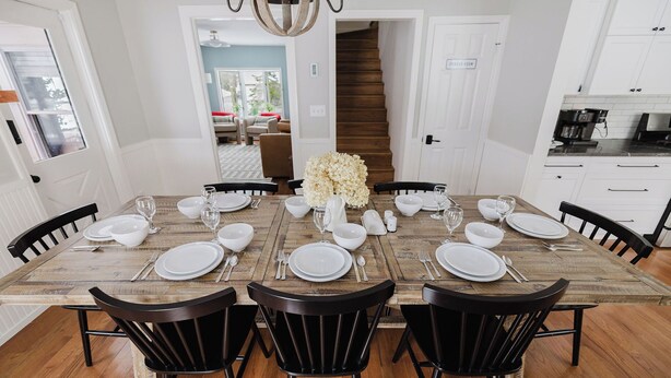 Un comedor con una mesa de madera preparada para seis comensales, rodeada de sillas negras. Un candelabro cuelga del techo, y se observa una escalera al fondo.