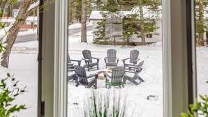 Vista desde una ventana a un espacio exterior cubierto de nieve, con sillas Adirondack dispuestas alrededor de una mesa.