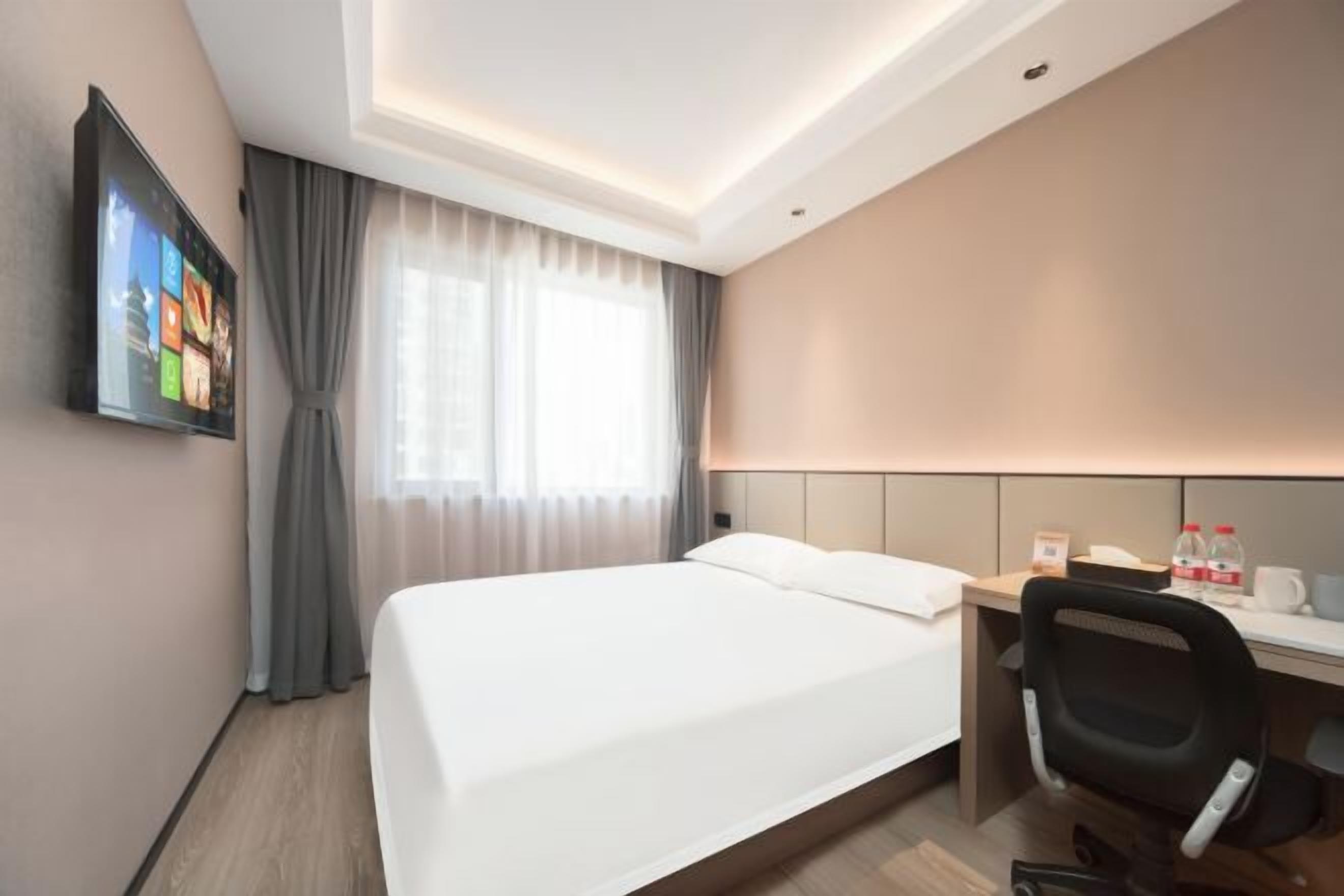 Quarto de hotel com uma cama, escrivaninha, cadeira, televisão na parede e uma janela com cortinas.