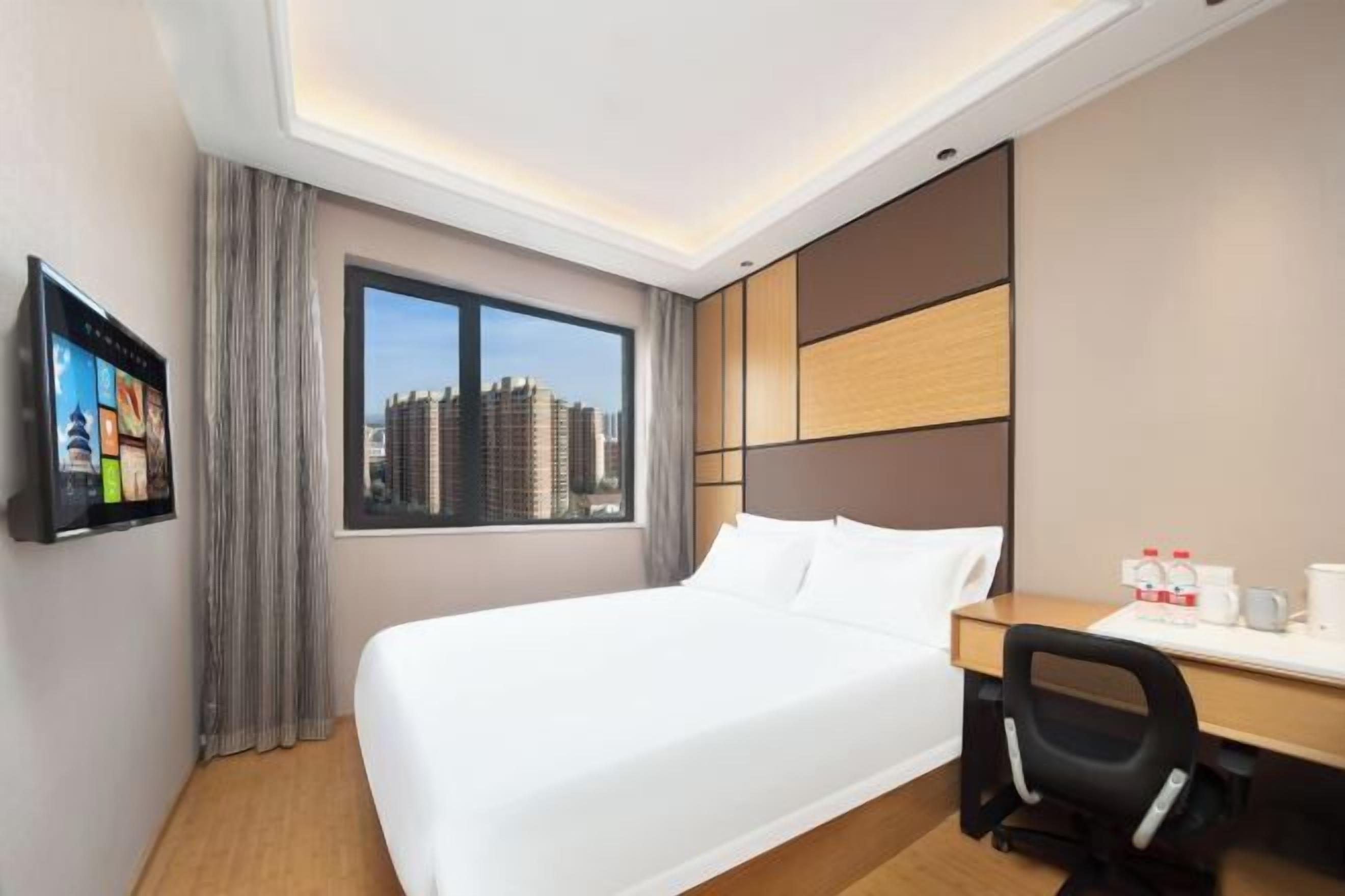 A hotel room with a large bed, a desk with a chair, a television, and a view of the cityscape.