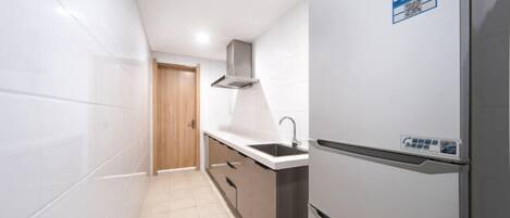 A narrow kitchen with white tiles, a stainless steel refrigerator, a wooden door, and a built-in oven.