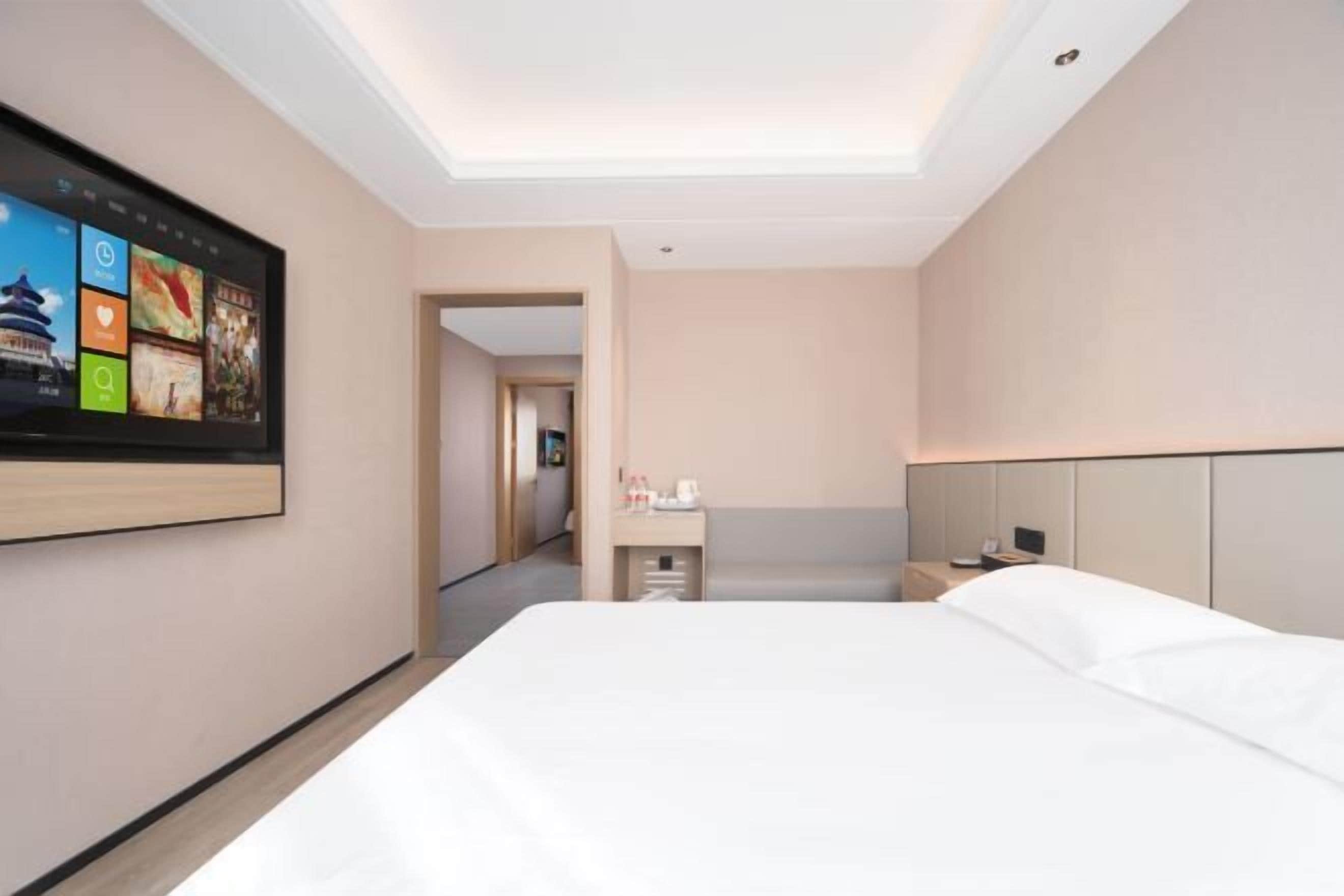 A hotel room with a large bed, a flat-screen TV mounted on the wall, a bedside table with a lamp, and a neutral color scheme.