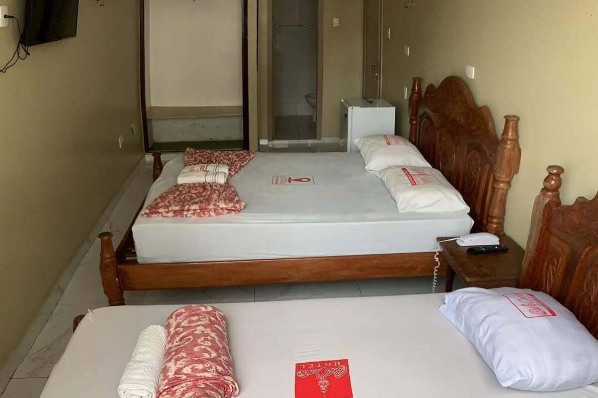 A hotel room with two wooden beds, each with a white mattress, red and white patterned blankets, and a red tag. There is a wooden nightstand with a phone and remote control, a small white table, and a wall-mounted TV.