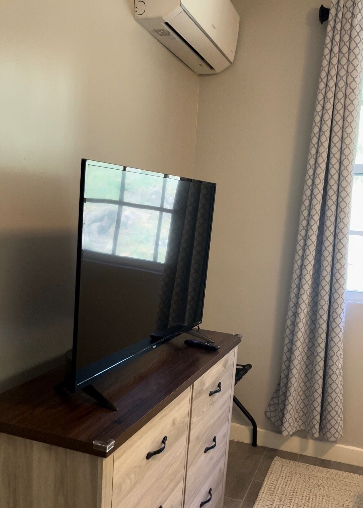 A flat-screen TV on a wooden cabinet, a window with a view, and a patterned curtain.