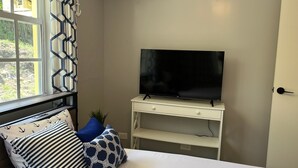 Un dormitorio con cama, televisor sobre un soporte blanco, ventana con cortinas y una puerta.