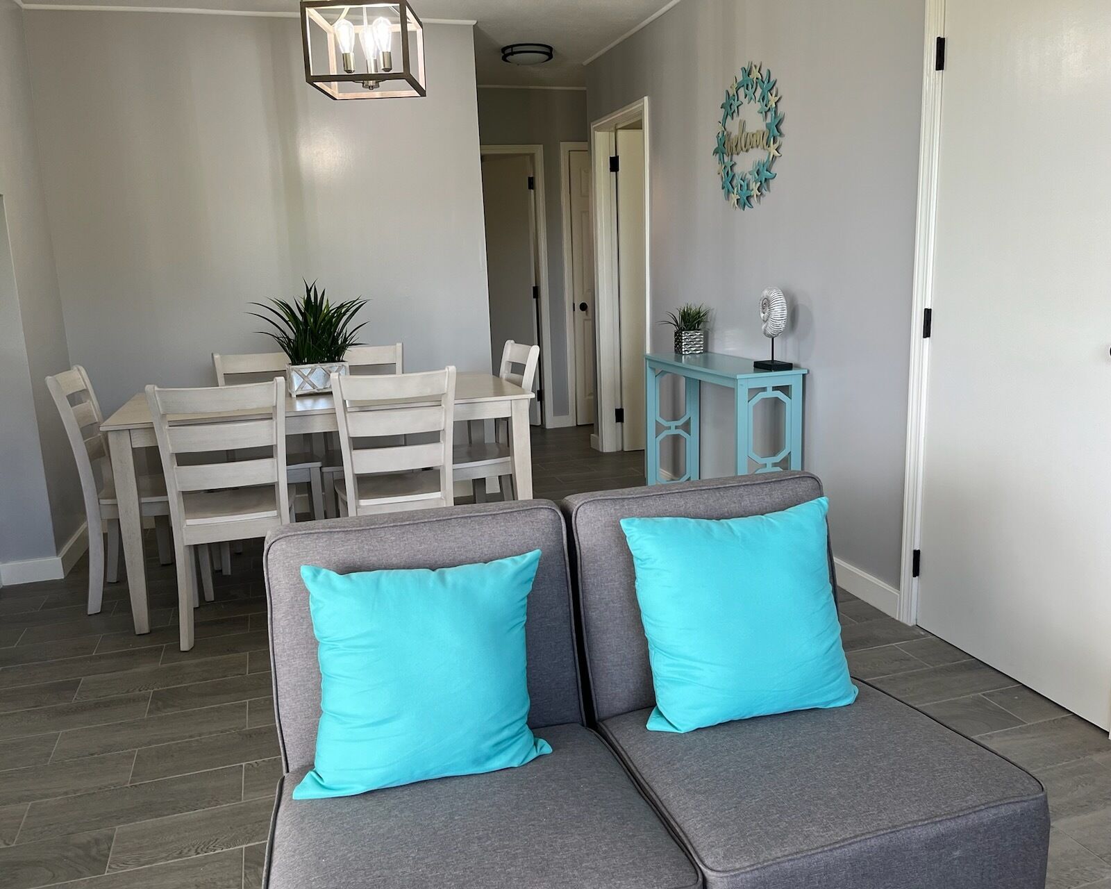 A modern living room with a gray sectional sofa, two turquoise pillows, a dining area with white chairs and a table, and a small blue side table.