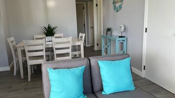 A modern living room with a gray sectional sofa, two turquoise pillows, a dining area with white chairs and a table, and a small blue side table.