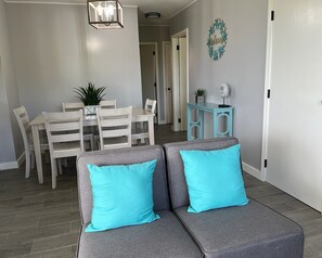 Un salon moderne avec un canapé d’angle gris, deux coussins turquoise, un coin repas avec des chaises blanches et une table, ainsi qu’une petite table d’appoint bleue.