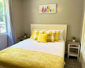 Un lit bien fait, avec une couverture jaune, une tête de lit blanche et des oreillers assortis. Un tableau représentant des voiliers est accroché au mur au-dessus du lit.