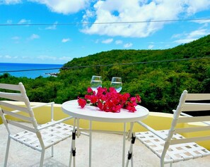 Une table ronde, deux chaises blanches, un vase de fleurs rouges et deux verres à vin. La table est placée devant un paysage verdoyant, avec une colline luxuriante en arrière-plan et un ciel bleu dégagé.