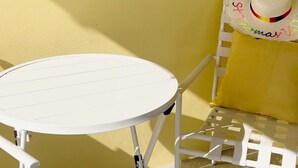 Un juego de mesa y silla de metal blanco frente a una pared amarilla, con un sombrero de paja que tiene texto colorido en el respaldo.