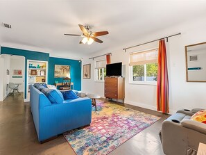 A modern living room with a blue sofa, a colorful rug, a ceiling fan, and a TV on a wooden cabinet. - Gulfport Getaway — Artsy Retreat 10 Min to Beach (Gulfport)